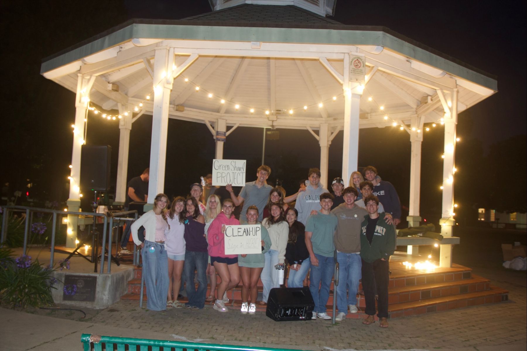 Green Shovel Project volunteers posing at night in front of a gazebo strung with lights, holding Green Shovel Project and clean-up sign-in signs